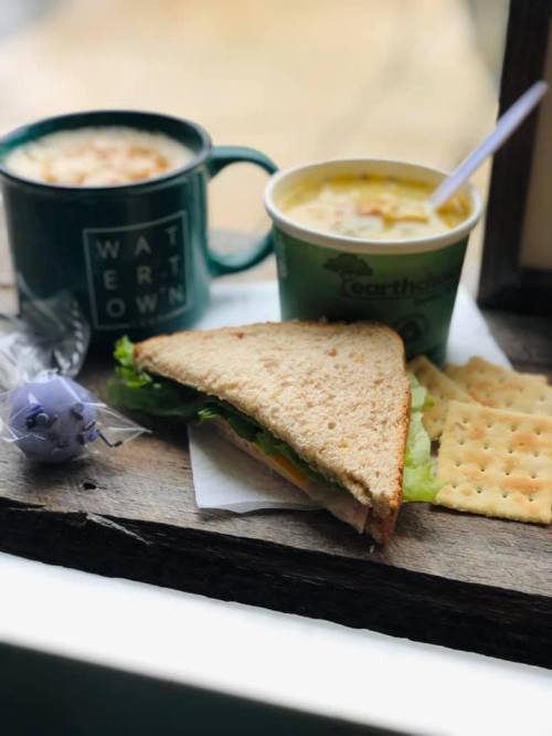 A sandwich with lettuce and cheese, a cup of soup, a mug, and crackers on a wooden surface by a window.