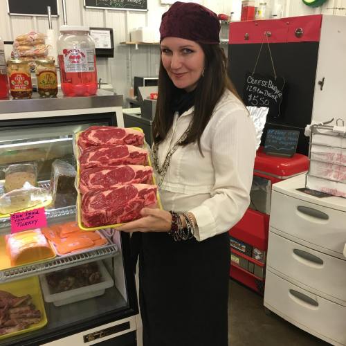 A woman in a butcher shop holds a tray of ribeye steaks, smiling at the camera with a vintage-style backdrop.