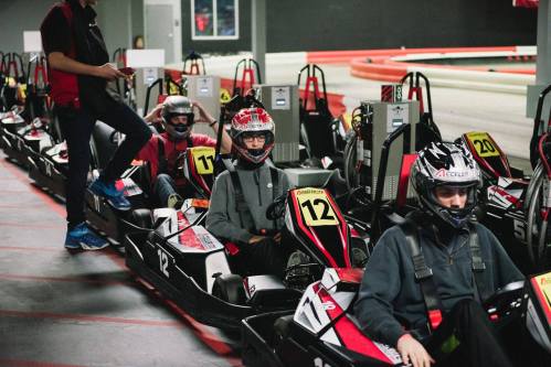 A group of go-kart racers in helmets, seated in karts, preparing for a race at an indoor track.