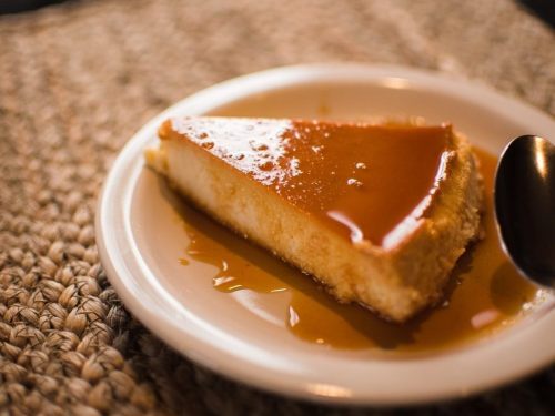 A slice of creamy flan on a white plate, drizzled with caramel sauce, resting on a textured surface.