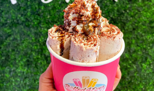 A hand holding a pink cup filled with rolled ice cream topped with whipped cream and chocolate sauce.
