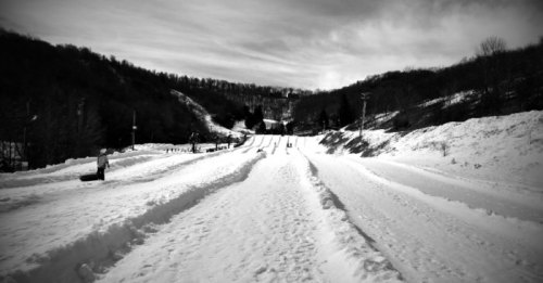 A snowy landscape with sledding tracks, surrounded by trees and hills under a cloudy sky.