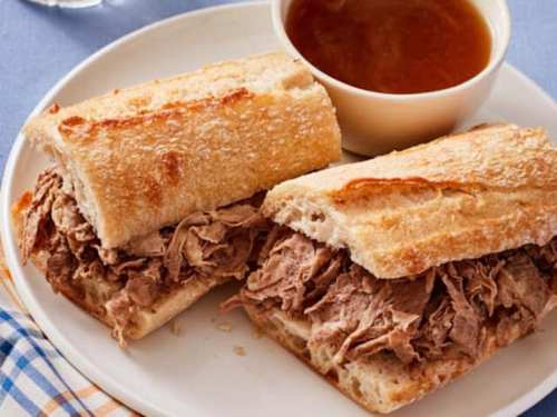 A plate with a sliced sandwich filled with shredded beef, served with a small bowl of dipping sauce.