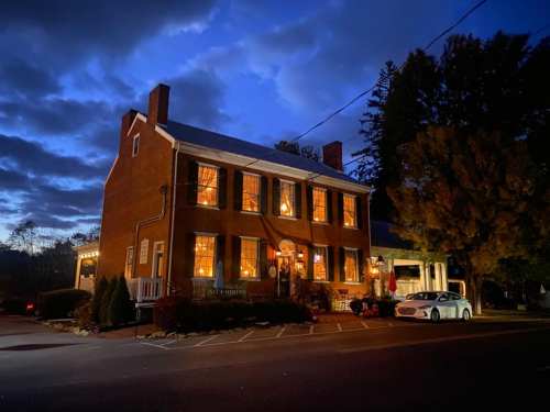 A charming brick building illuminated at dusk, with large windows glowing warmly against a cloudy sky.