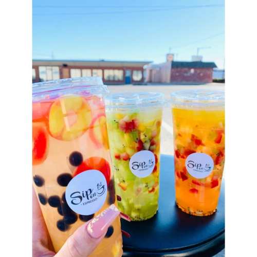 Three colorful fruit drinks in clear cups with lids, featuring vibrant fruits and a logo, set against a sunny background.