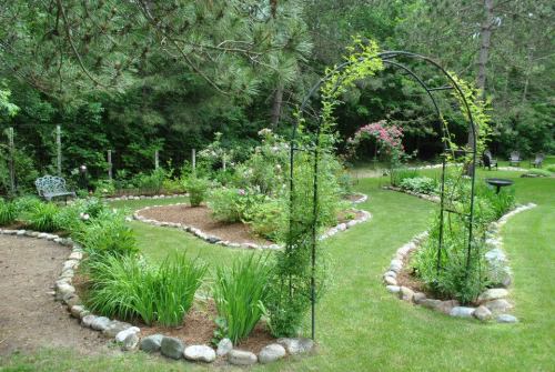 A lush garden with curved pathways, flower beds, and a metal archway surrounded by greenery and trees.