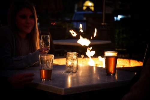 A woman sits at a table by a fire pit, enjoying drinks with a warm glow from the flames in the evening.
