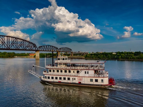 A riverboat cruises on a calm river under a blue sky with fluffy clouds, near a bridge in the background.