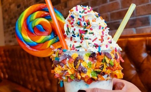 A colorful milkshake topped with whipped cream, sprinkles, and a large rainbow lollipop, held against a brick wall.