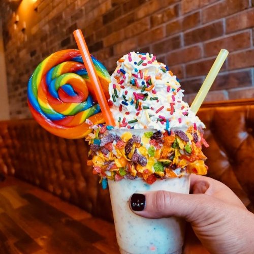 A colorful milkshake topped with whipped cream, sprinkles, and a large rainbow lollipop, held against a brick wall.
