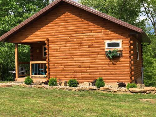 A rustic log cabin with a porch, surrounded by greenery and landscaped stones.