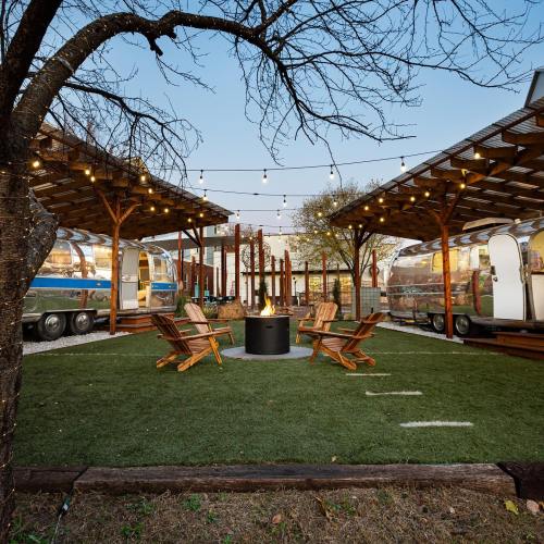 A cozy outdoor space with vintage Airstreams, wooden seating, and string lights around a fire pit on green grass.