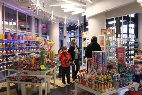 A colorful candy store filled with various sweets, with customers browsing and chatting among vibrant displays.