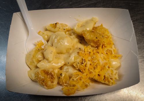 A serving of creamy macaroni and cheese topped with crispy cheese and breadcrumbs in a paper dish.