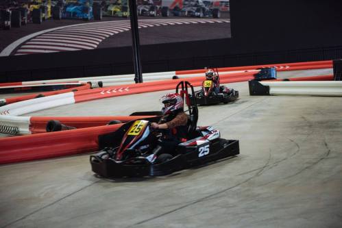 Two go-karts racing on an indoor track, with one kart in the foreground and a colorful backdrop of racing cars.
