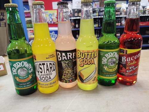 A lineup of six unique soda bottles: ginger ale, mustard, barf, butter, and bacon flavors, displayed on a counter.