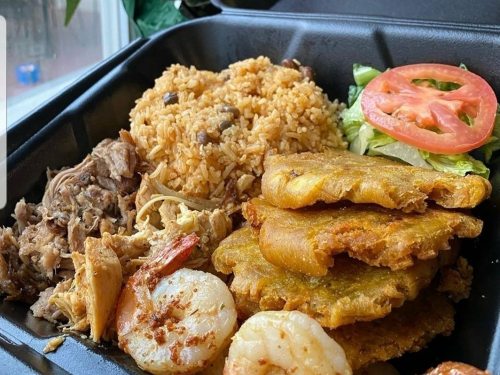 A takeout container filled with rice, shredded chicken, shrimp, fried plantains, and a side of lettuce and tomato.