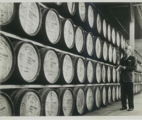 A man in a suit stands beside rows of stacked barrels in a warehouse, inspecting the labels on the barrels.