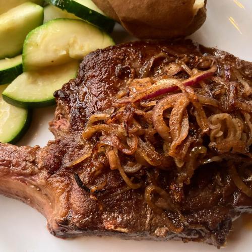 A juicy grilled pork chop topped with caramelized onions, served with sliced cucumbers and a baked potato.
