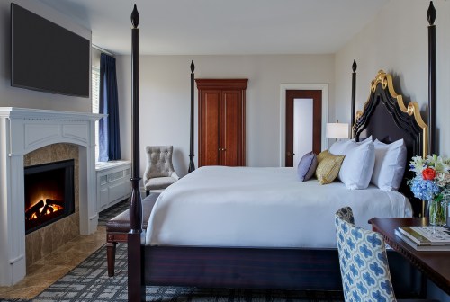A stylish hotel room featuring a king bed, fireplace, armchair, and a desk with flowers, all in a modern design.