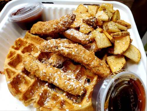 A takeout container with a waffle topped with fried chicken strips, seasoned potato wedges, and two dipping sauces.