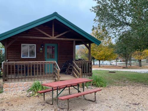 A cozy log cabin with a green roof, porch, and picnic table, surrounded by trees and a gravel path.