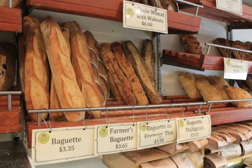 A display of various types of bread, including baguettes and whole wheat, with price tags on a wooden shelf.