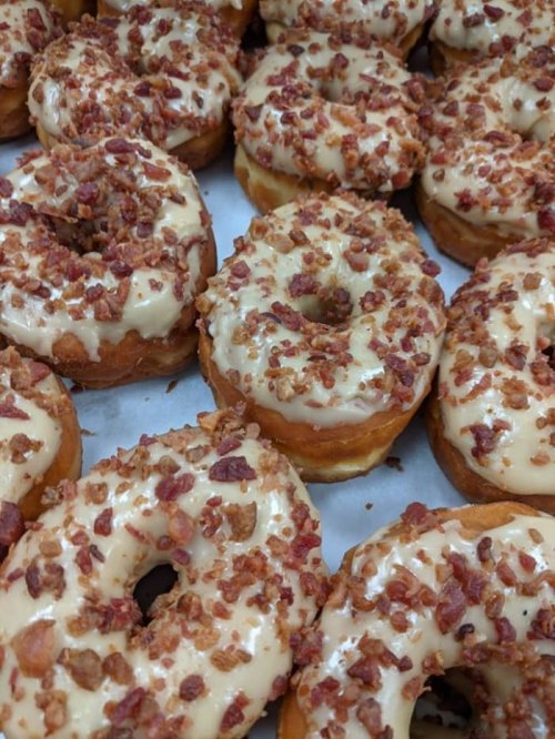 A close-up of glazed donuts topped with crispy bacon bits, arranged neatly on a tray.