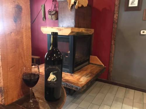 A bottle of red wine and a glass sit on a wooden table near a cozy fireplace in a rustic setting.