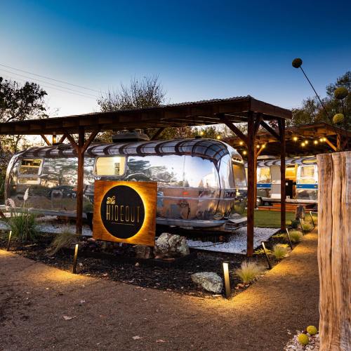 A shiny airstream trailer under a wooden structure, illuminated by warm lights, with a sign reading "The Hideout."