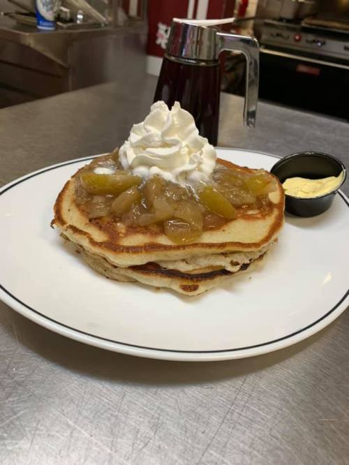 A stack of pancakes topped with whipped cream and fruit compote, served with a side of butter and syrup.
