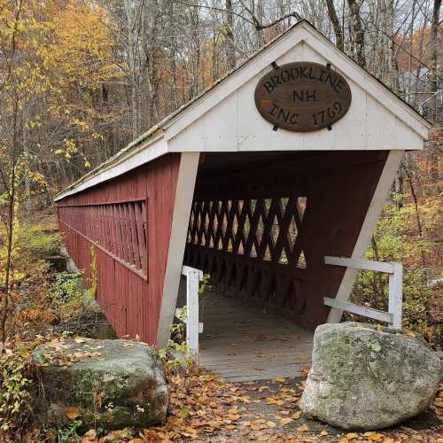 A red covered bridge in Brookline, NH, surrounded by autumn foliage and stone features. Sign reads "Brookline NH, Inc. 1769."