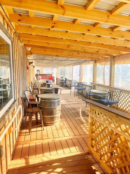 A wooden deck with tables and chairs, featuring a barrel and a covered area, surrounded by a rustic wooden structure.