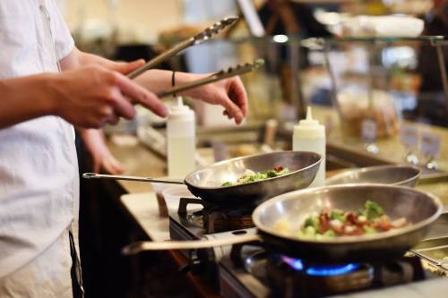 A chef uses tongs to stir vegetables in two pans on a stove in a busy kitchen.