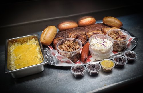 A platter of barbecue with pulled pork, sausage, coleslaw, cheesy rice, and rolls, accompanied by various sauces.