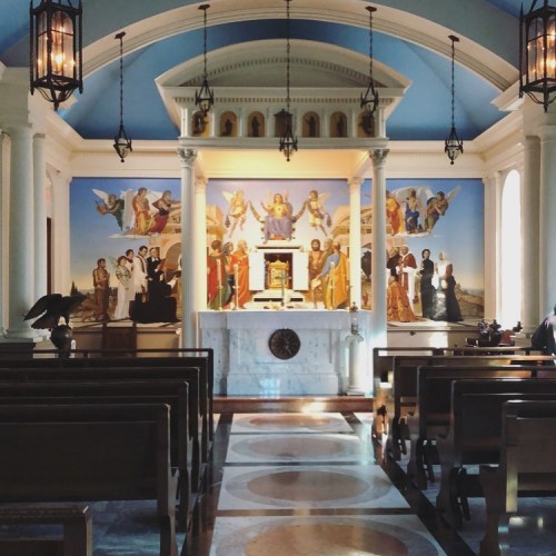 Interior of a church featuring a colorful mural, altar, and wooden pews under a blue ceiling with hanging lanterns.