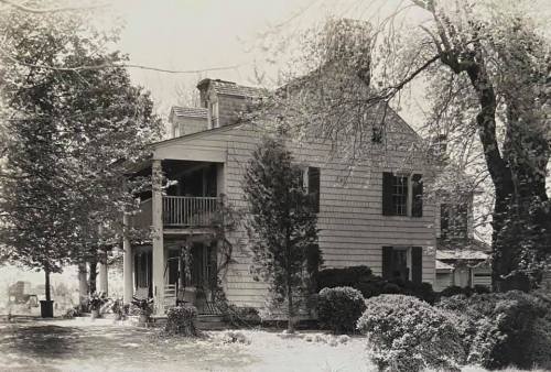 A vintage black-and-white photo of a two-story house surrounded by trees and shrubs.