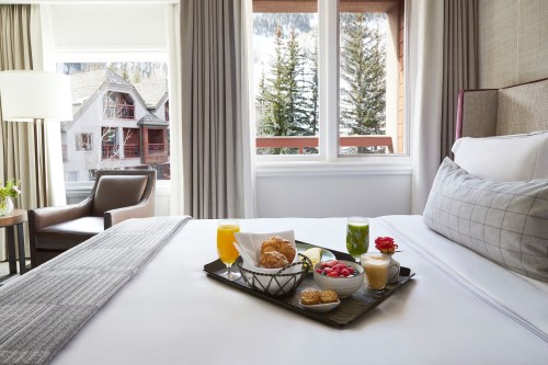 A cozy hotel room with a bed and a tray of breakfast items, including drinks, pastries, and fruit, by a window.