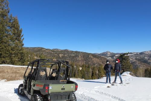 Two people stand on snow-covered terrain with mountains in the background, next to an off-road vehicle. Clear blue sky above.