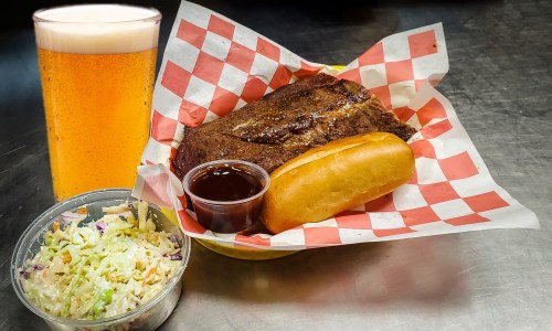 A plate with barbecue meat, a bun, coleslaw, and a glass of beer on a metal surface.