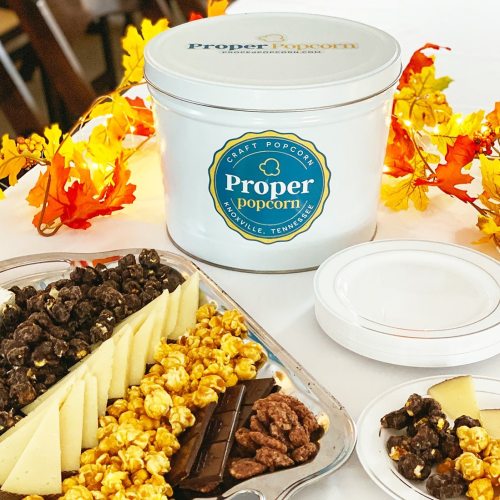 A white container of Proper Popcorn surrounded by popcorn varieties, cheese, chocolate, and autumn leaves.