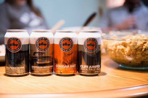 Four cans of Country Boy Brewing beer lined up on a table, with a bowl of food in the background.