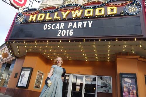 A woman in a vintage dress stands in front of a theater marquee announcing an Oscar Party in 2016.