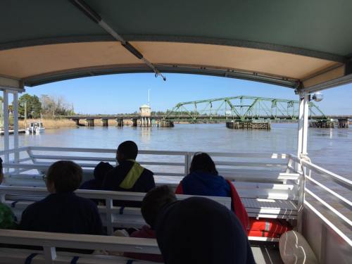View from inside a boat showing a green bridge over a river, with passengers seated in the foreground.