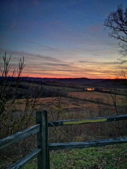A serene landscape at sunset, featuring a wooden fence, rolling hills, and a colorful sky with hints of water in the distance.