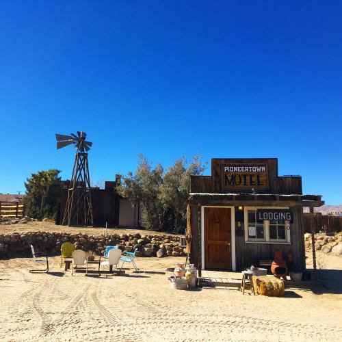 A rustic motel in Pioneertown with a windmill, surrounded by desert landscape and vintage chairs in front.