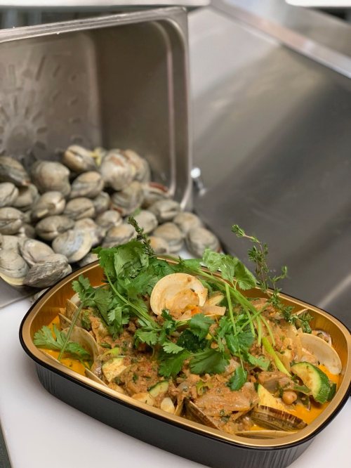 A dish of seafood stew with clams, herbs, and spices, next to a pile of fresh clams in a kitchen setting.