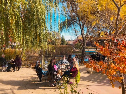 A cozy outdoor scene with people sitting around tables, surrounded by trees and autumn foliage near a pond.
