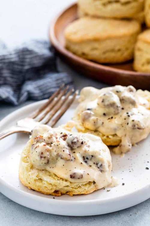 Two biscuits topped with creamy sausage gravy, served on a white plate with a fork, and a wooden bowl of biscuits in the background.
