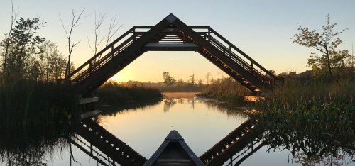 A wooden bridge arches over a calm waterway at sunrise, reflecting in the water surrounded by lush greenery.